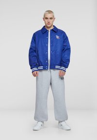Niebieska satynowa kurtka w stylu varsity z białymi paskami na mankietach, z wyszytym logo oraz szare dresy z nadrukowanym tekstem. Białe sneakersy.