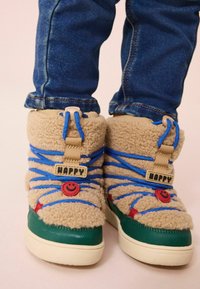 Bottes chevilles moelleuses et brunes avec des lacets bleus ; elles présentent des accents sourire rouges et une étiquette « HAPPY » ; semelles en caoutchouc vertes ; matériau extérieur texturé.