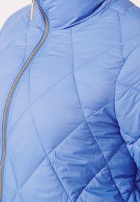 Giacca trapuntata light blue con colletto alto, dotata di zip dorata e motivo a diamante. Esterno liscio e lucido con sezioni imbottite.