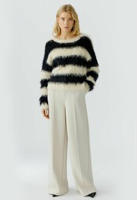 Schwarz- und cremefarbig gestreifter, flauschiger Pullover mit langen Ärmeln, kombiniert mit hoch geschnittenen, weiten cremefarbenen Hosen und spitzen schwarzen Absatzschuhen.