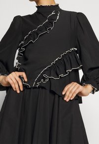 Svart blus med volangdetaljer och vit fåll, med hög hals och långärmade, tillverkad av texturerad tyg.