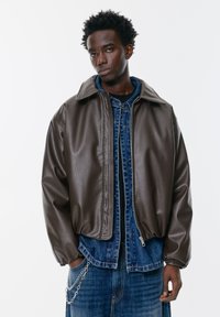 Hnedá kožená bomber bunda s golierom, so zipsovým zapínaním a textúrovaným povrchom, prekrytá modrou džínovou bundou. Reťazový detail na vrecku.