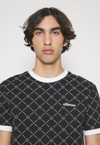 Black t-shirt with white geometric pattern, ribbed white collar, and a small black "ellesse" logo on the chest. Soft cotton material.
