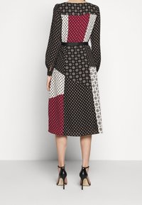 Žena v černých midi šatech s dlouhými rukávy, které mají patchworkový vzor s červenými, bílými a béžovými geometrickými potisky, v černých vysokých podpatcích.