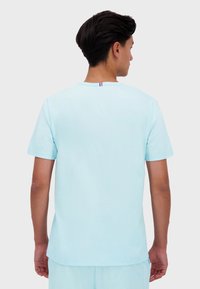 Světle modré tričko s krátkým rukávem vyrobené z měkké tkaniny, s kulatým výstřihem a malým červeným, bílým a modrým detailem na zadní straně.