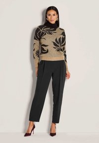 Frau in beigem und schwarzem Blumensweater, schwarzen hoch taillierten Hosen und dunkelroten High Heels, stehend vor einer hellen Wand.