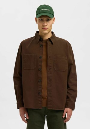 Joven con una gorra verde, chaqueta marrón de botones sobre una camisa marrón y pantalones verde oscuro, de pie contra un fondo blanco liso.
