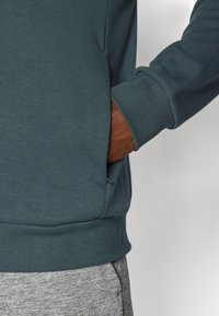 Kokvilnas jaukta auduma džemperis bālganā teal krāsā ar ribinātām aprocēm un jostu. Tam ir kabata kreisajā pusē, savienots ar pelēkiem teksturētiem biksēm.