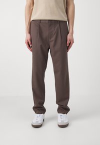 Un pantalon marron en tissu lisse avec une coupe décontractée, comportant des plis avant, des passants de ceinture et des poches latérales, porté avec des baskets blanches.