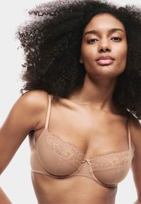 Beige bh med blonderkant og tekstureret stof samt en lille sløjfe i midten, med justerbare stropper og en struktureret form.