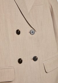 Blazer beige à double boutonnage en tissu texturé, avec des boutons noirs et une poche poitrine.