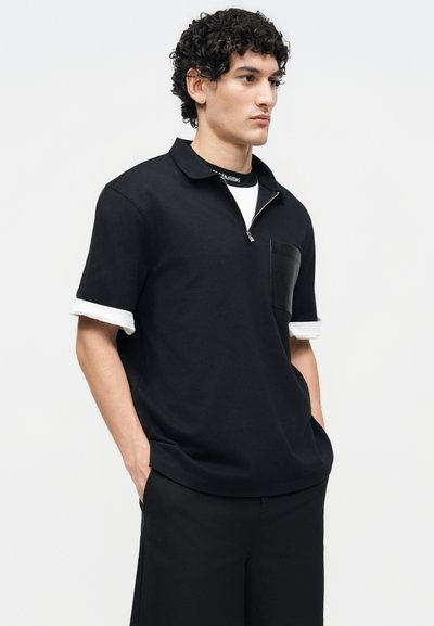 Νέος άνδρας με σγουρά μαλλιά που φοράει μαύρο κοντομάνικο πουκάμισο με φερμουάρ στον γιακά πάνω από ένα λευκό φανελάκι, χέρια στις τσέπες μαύρου παντελονιού, κοιτάζει πλάγια.