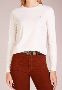 Långärmad ljusrosa bomullsskjorta med en liten blå logotyp på bröstet, i kombination med rostfärgade kordbyxor och ett svart bälte.