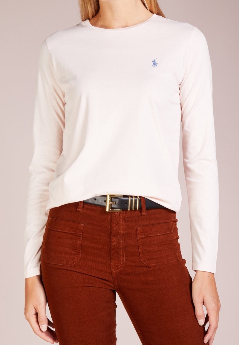 Långärmad ljusrosa bomullsskjorta med en liten blå logotyp på bröstet, i kombination med rostfärgade kordbyxor och ett svart bälte.