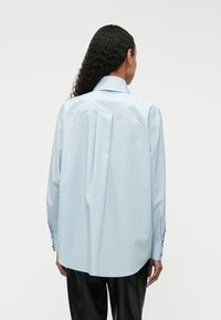 Γαλάζιο πουκάμισο με κουμπιά, με μυτερό γιακά, μακριά μανίκια και πτυχές στην πλάτη; κατασκευασμένο από λεία, ελαφριά ύφανση.