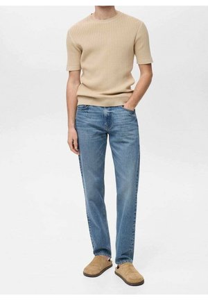 Mand iført beige ribbet kortærmet sweater, blå straight-leg jeans og brune slip-on sko, stående med den ene hånd i lommen.