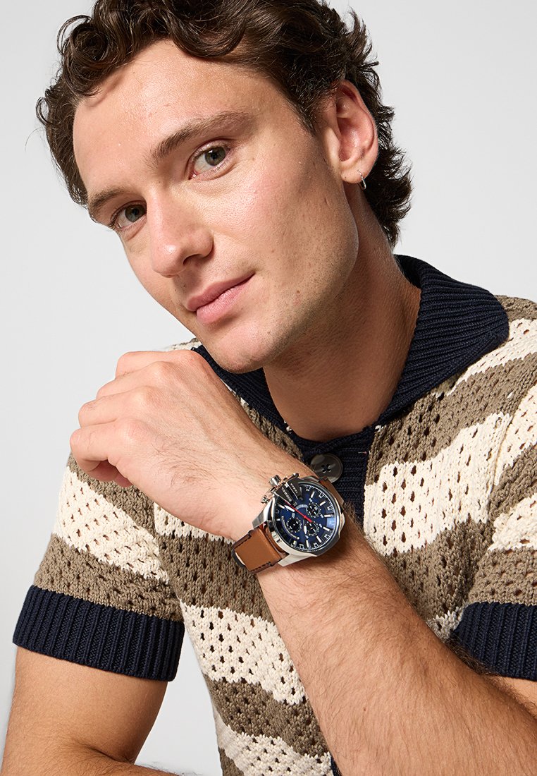 Chronograph-Uhr mit blauem Zifferblatt, silbernem Metallgehäuse und braunem Lederarmband. Strickpullover in beige und braunen Streifen mit marineblauen Akzenten.