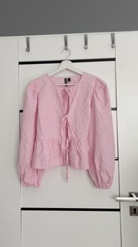 Blusa rosa chiaro con righe verticali, scollatura rotonda, maniche lunghe a sbuffo, chiusura con fiocco davanti e orlo a peplo. Tessuto testurizzato.