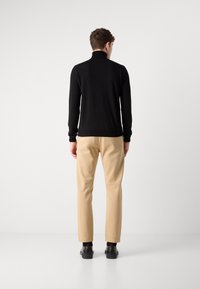 Homme debout de dos, portant un pull à col roulé noir, un pantalon beige, des chaussettes noires et des chaussures noires sur un fond blanc uni.