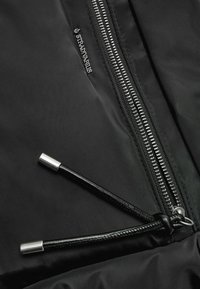 Detail einer schwarzen Steppjacke mit einem silbernen Reißverschluss, schwarzem Kordelzug mit silbernen Enden und einem dezenten, in den Stoff eingravierten Logo.
