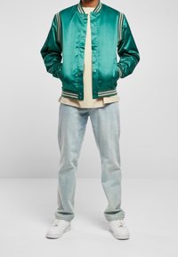 Grüne Satin-Bomberjacke mit gestreiften Akzenten, Knopfverschluss und zwei Seitentaschen, kombiniert mit einem beigen Hemd und hellen Jeans aus Denim.