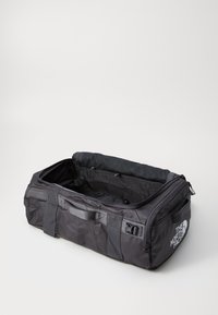 Black nylon duffel bag with two top handles, zip closure, mesh pocket, and side buckles. Features a white logo on one side.