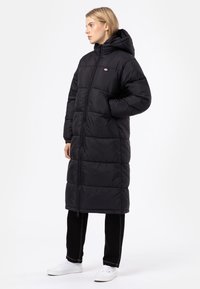 Long black puffer coat with a hood, featuring a front zipper and stitched square patterns. Worn over black pants and white sneakers.