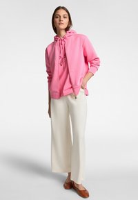 Felpa rosa con zip, con coulisse, tasca frontale e dettagli del logo; abbinata a pantaloni bianchi larghi e scarpe slip-on marroni.