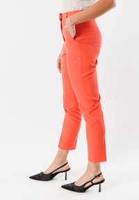 Pantaloni attillati corallo con tasche laterali, design affusolato e vita media, abbinati a scarpe slingback nere con punta.