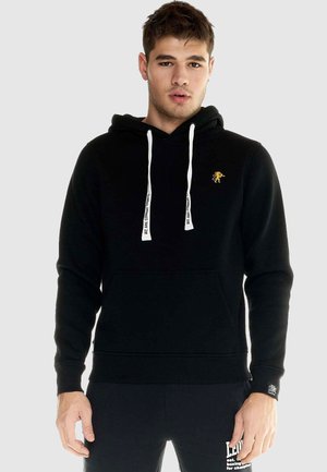 Homme portant un sweat à capuche noir avec cordons blancs et petit emblème de lion jaune, associé à un pantalon noir avec texte blanc sur la jambe.