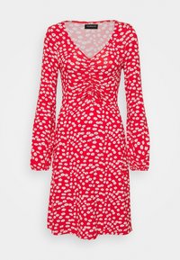 Czerwona sukienka z długimi rękawami, dekoltem w kształcie litery V, z białymi wzorami w stokrotki, marszczonym przodem oraz luźnym krojem. Miękki, lekki materiał.