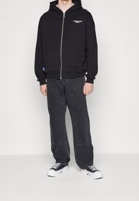 Black zip-up hoodie with front pockets, worn with dark gray wide-leg jeans and black sneakers. Notable white printed logo on the chest.