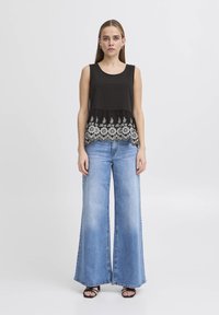 Top senza maniche nero con un orlo ricamato bianco simile a pizzo, abbinato a jeans in denim azzurro a gamba larga e sandali neri.