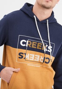 Sudadera con capucha en azul marino y mostaza, con un bolsillo frontal tipo canguro y un diseño gráfico en el texto. Material de mezcla de algodón, ajuste casual.