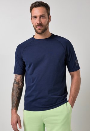 Mand med skæg og tatoveret venstre arm bærer navy kortærmet skjorte og lysegrønne bukser, stående mod en ensfarvet grå baggrund.