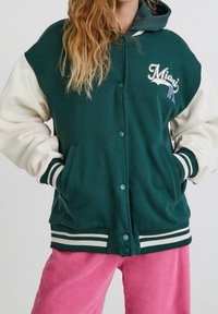 Chaqueta universitaria verde y crema con capucha, botones a presión, bolsillos delanteros y bordado "Miami". Puños y dobladillo de canalé a rayas.