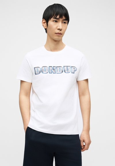 Νέος άνδρας με μαύρα μαλλιά, φορώντας λευκό T-shirt με το λογότυπο "DONDUP" και σκοτεινό παντελόνι, στέκεται με τα χέρια στις τσέπες μπροστά σε απλό φόντο.