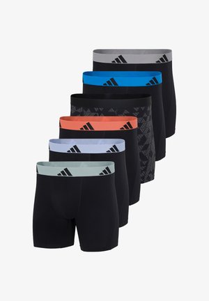 Athletischer Mann steht barfuß, trägt schwarze Boxer-Shorts mit einem orangenem Bund, der schwarze Adidas-Logos zeigt.