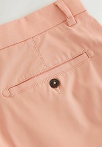 Gros plan sur un pantalon rose clair montrant une poche arrière passepoilée avec un seul bouton foncé et une boucle de ceinture sur la taille.