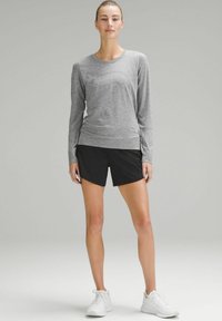Grijs shirt met lange mouwen gecombineerd met zwarte shorts, met een gladde textuur. De outfit wordt afgewerkt met witte sportschoenen.