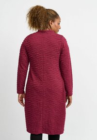 Burgunderfarbendes, strukturiertes Kleid mit gewellten Linien, langen Ärmeln und hohem Kragen, getragen mit schwarzen Leggings. Rückansicht gezeigt.