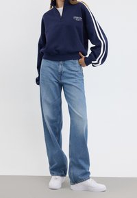 Sudadera polar azul marino con medio cierre y rayas blancas en las mangas, combinada con vaqueros de pierna ancha azul claro y zapatillas blancas.