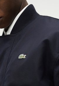 Gros plan sur une personne portant une veste bleu marine avec un col côtelé et un petit logo de crocodile vert brodé sur la poitrine.