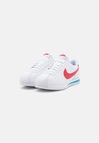 Hvide sportsko med en glat læderfinish, rød Nike-swoosh, lyserødt logo på tungen og en blå detalje på sålen.