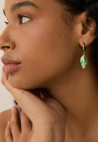 Grøn skjelsformet ørering med en guldring, hængende nær modellens øre. Glat tekstur og indviklet detaljering synlig.