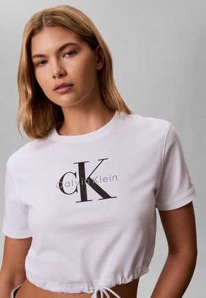Kvinne med skulderlangt blond hår iført en hvit cropped Calvin Klein t-skjorte med svart logo på grå bakgrunn.