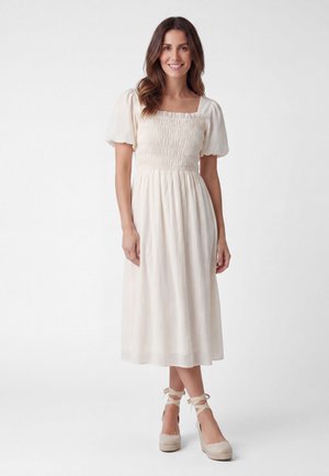 Vrouw met lang bruin haar die een witte smock midi-jurk draagt met pofmouwen en beige sleehak espadrilles met enkelbandjes.