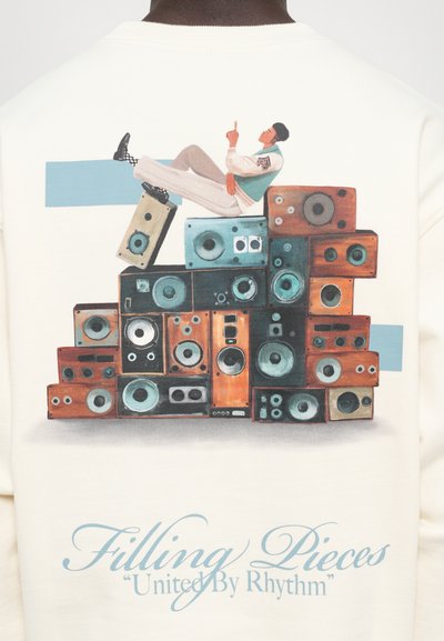 Pessoa reclinada sobre colunas vintage empilhadas com formas geométricas azuis e texto "Filling Pieces United By Rhythm" em tecido creme.