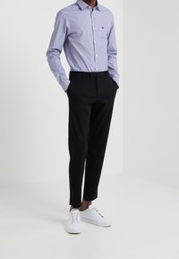 Chemise à boutons bleu rayée, pantalons noirs et baskets blanches. La chemise a un petit logo ; les pantalons sont ajustés avec une texture lisse.