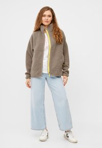Graue Fleecejacke mit hohem Kragen, gelbem Frontreißverschluss und seitlichen Taschen, kombiniert mit hellblauen weit geschnittenen Jeans und weißen Sneakers.